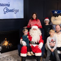 Little girl with green dress sits on Santa's lap, boy in gray sweater sits on dad's lap, while mom and other child stand by Louie